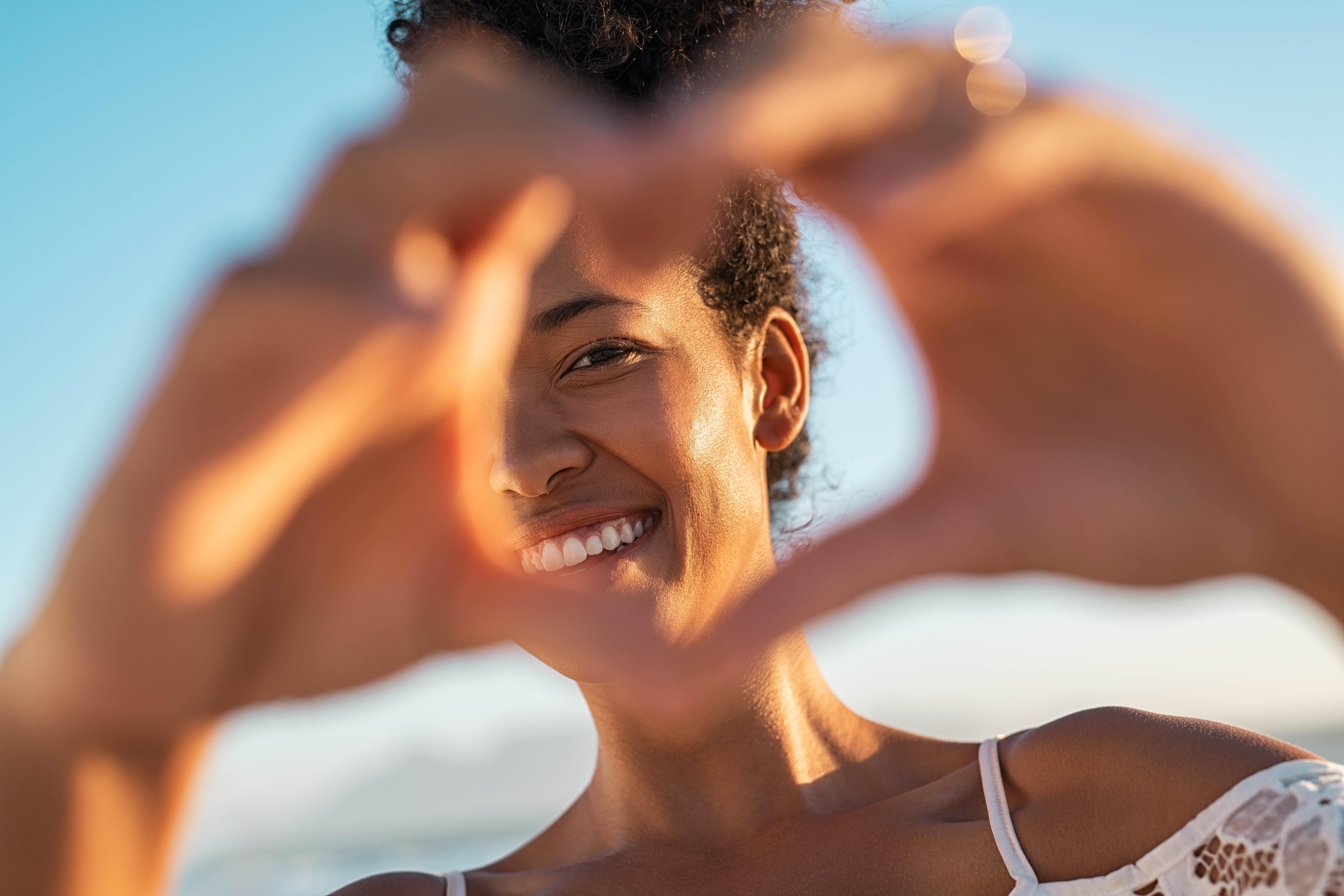 Young black woman with her hands in the shape of a heart in front of her face. Bright blue sky in the background